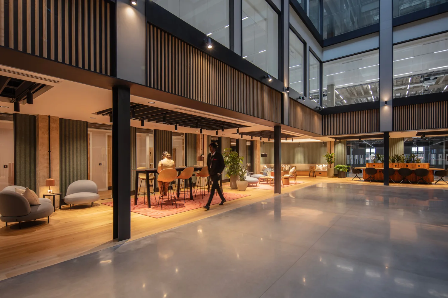 Modern open-plan office interior with double-height ceiling, glass-walled upper level, and wood slat panels. Lounge seating, tables, and plants arranged on rugs; one person seated at a table and another walking across polished floor.