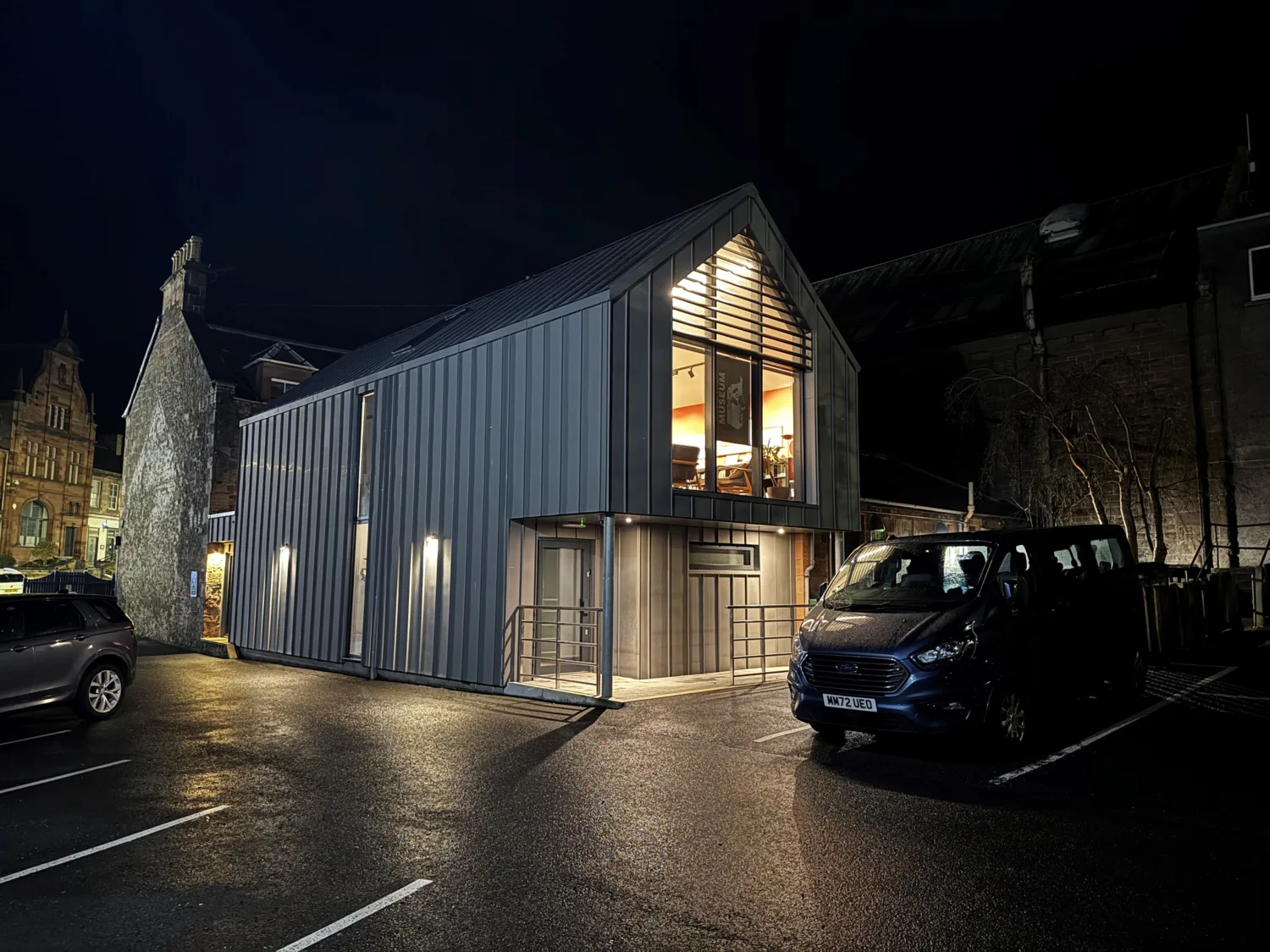 Modern two-storey building with a sloped metal roof and vertical panel siding, lit at night. Large upper window shows tables and chairs inside. Two vehicles parked on wet asphalt in front.
