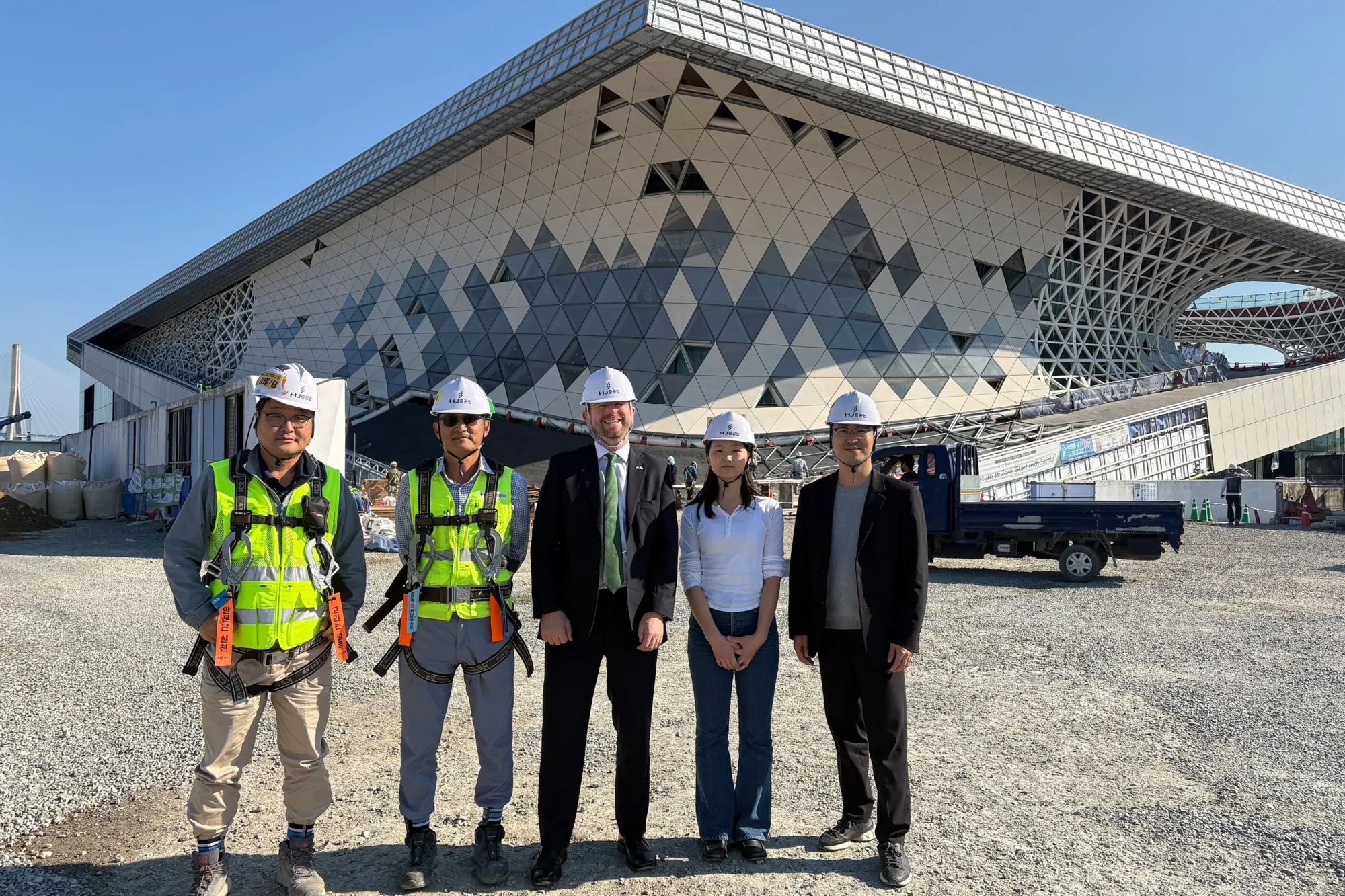 Five people wearing safety helmets stand outside a large geometric-clad building under construction with exposed white steel latticework.