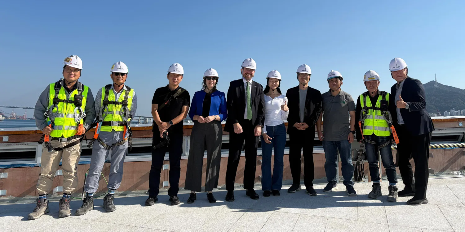 Ten people wearing safety helmets stand together on a construction site, with a waterfront, bridge and hillside in the background.