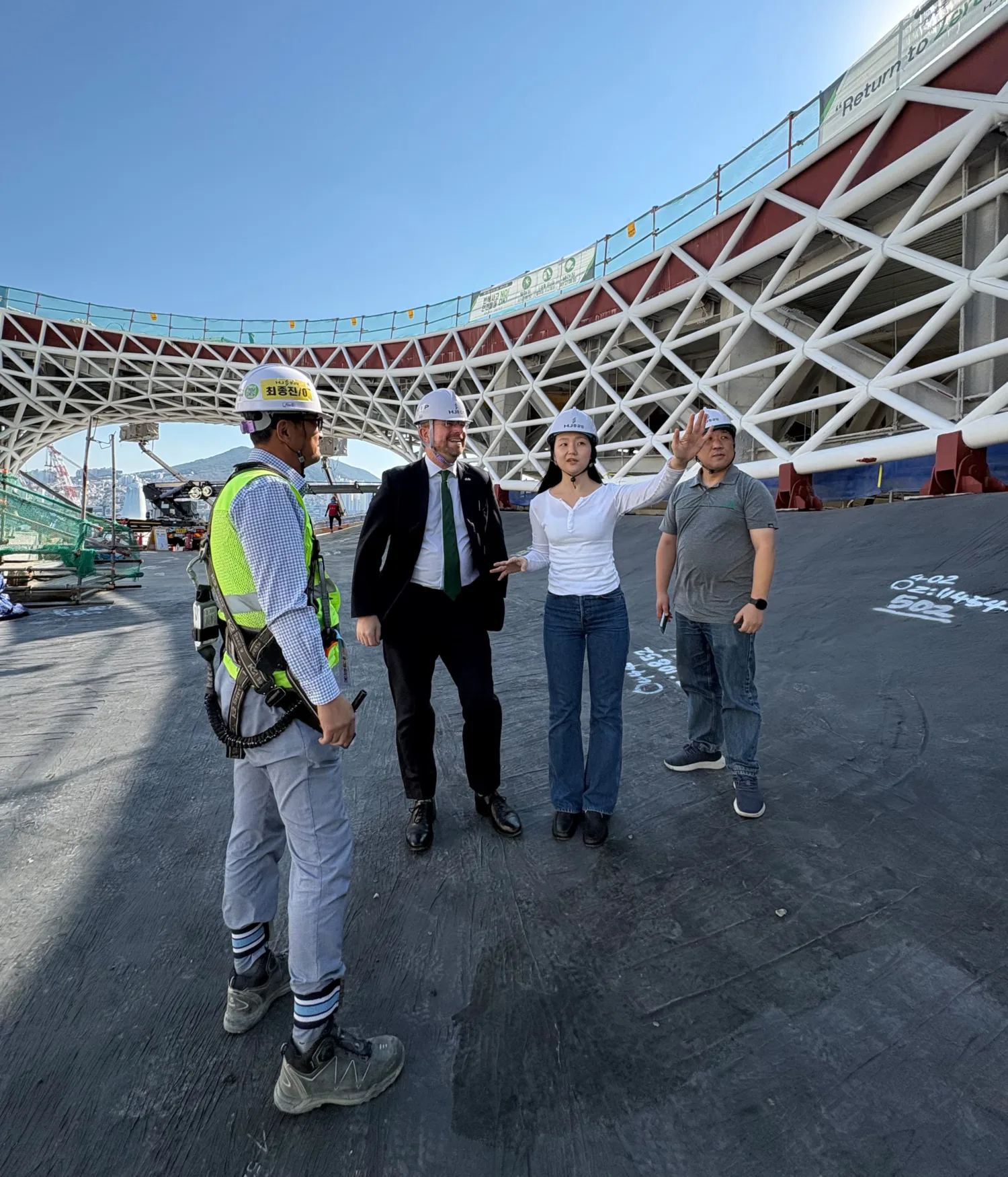 Four people wearing construction helmets stand beneath a large white steel lattice structure during a construction site visit.