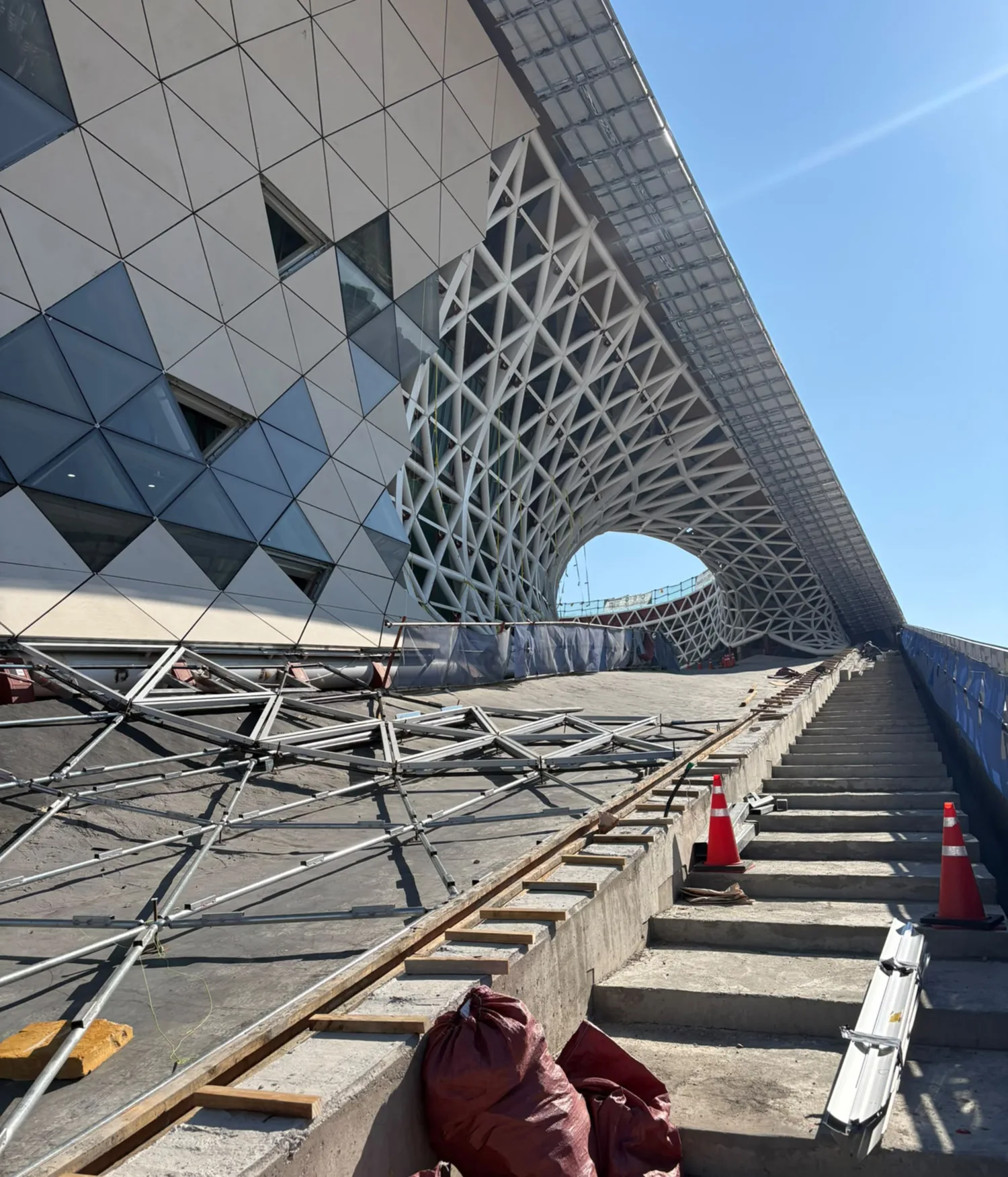 Triangular façade panels and exposed white steel latticework frame a curved exterior walkway under construction.