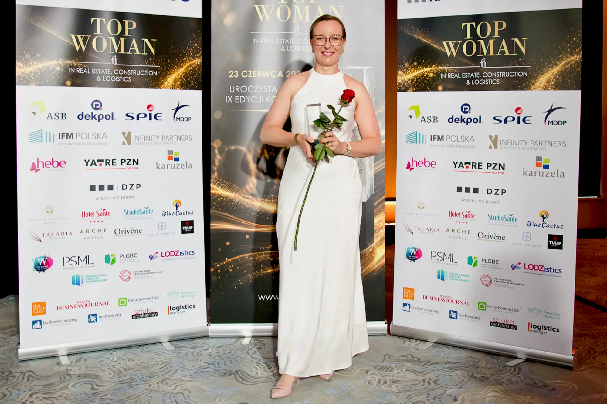 Person holding a glass award and a red rose while standing between event banners displaying sponsor logos and award branding.