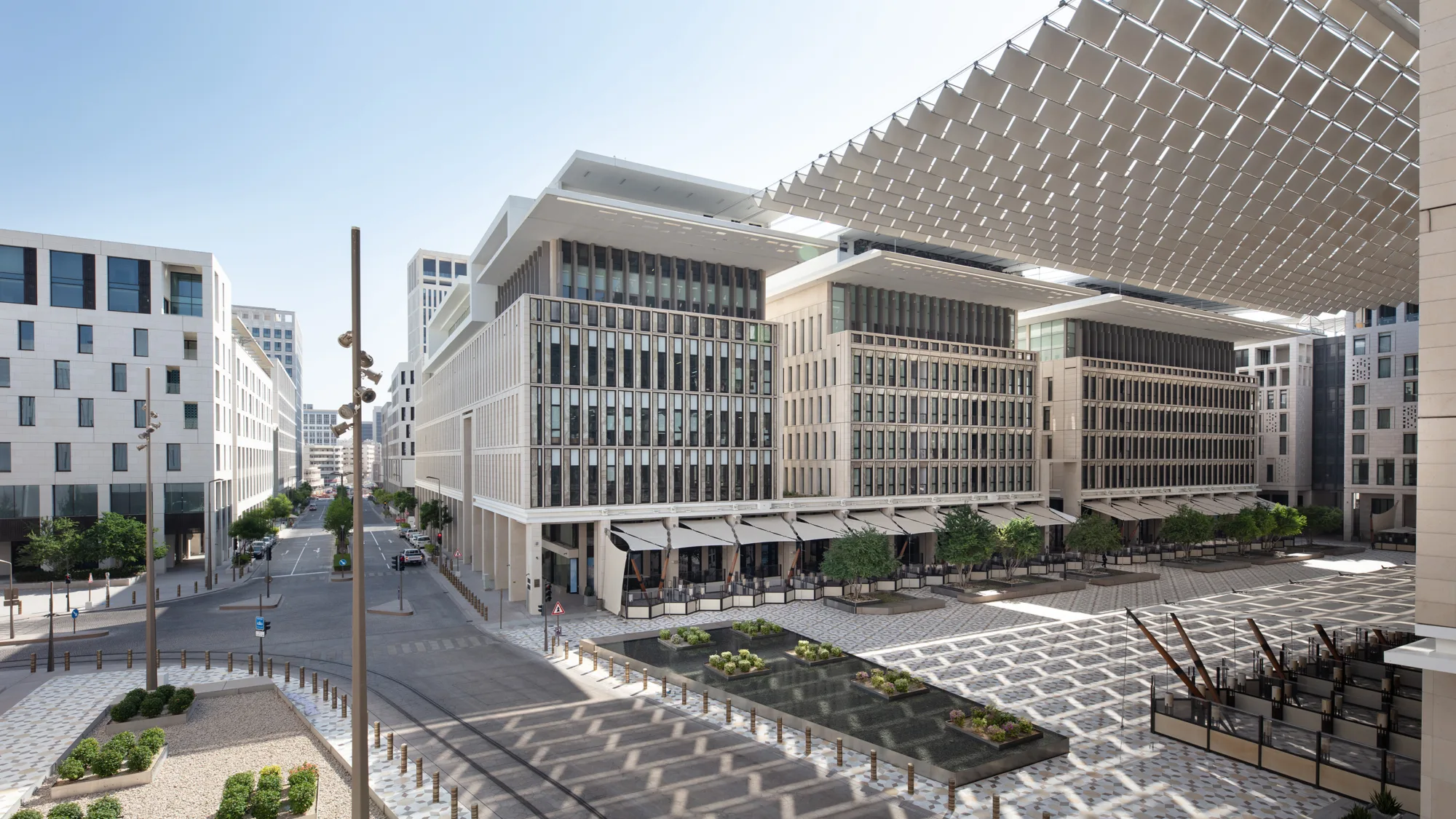 Modern urban plaza with mid-rise buildings featuring glass windows and white facades, patterned pavement, landscaped plant areas, and a large overhead shade structure. No visible text or people.