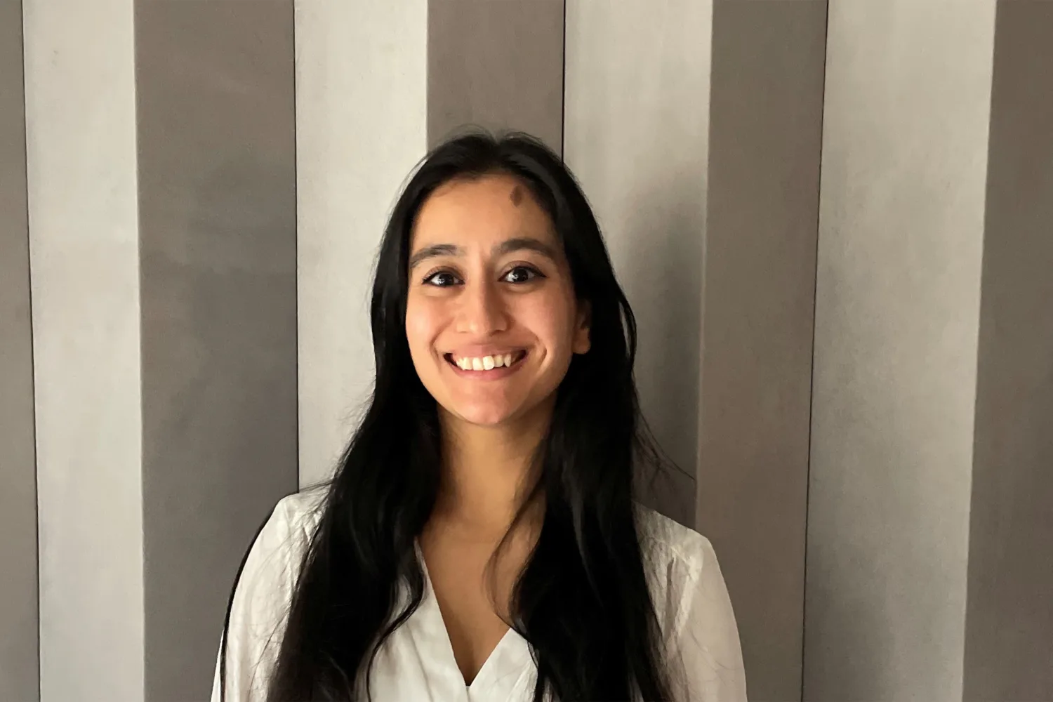 Head-and-shoulders portrait of a person with long dark hair wearing a white top, facing the camera with teeth visible. Background shows vertically striped wall panels in neutral tones; no visible text or additional objects.
