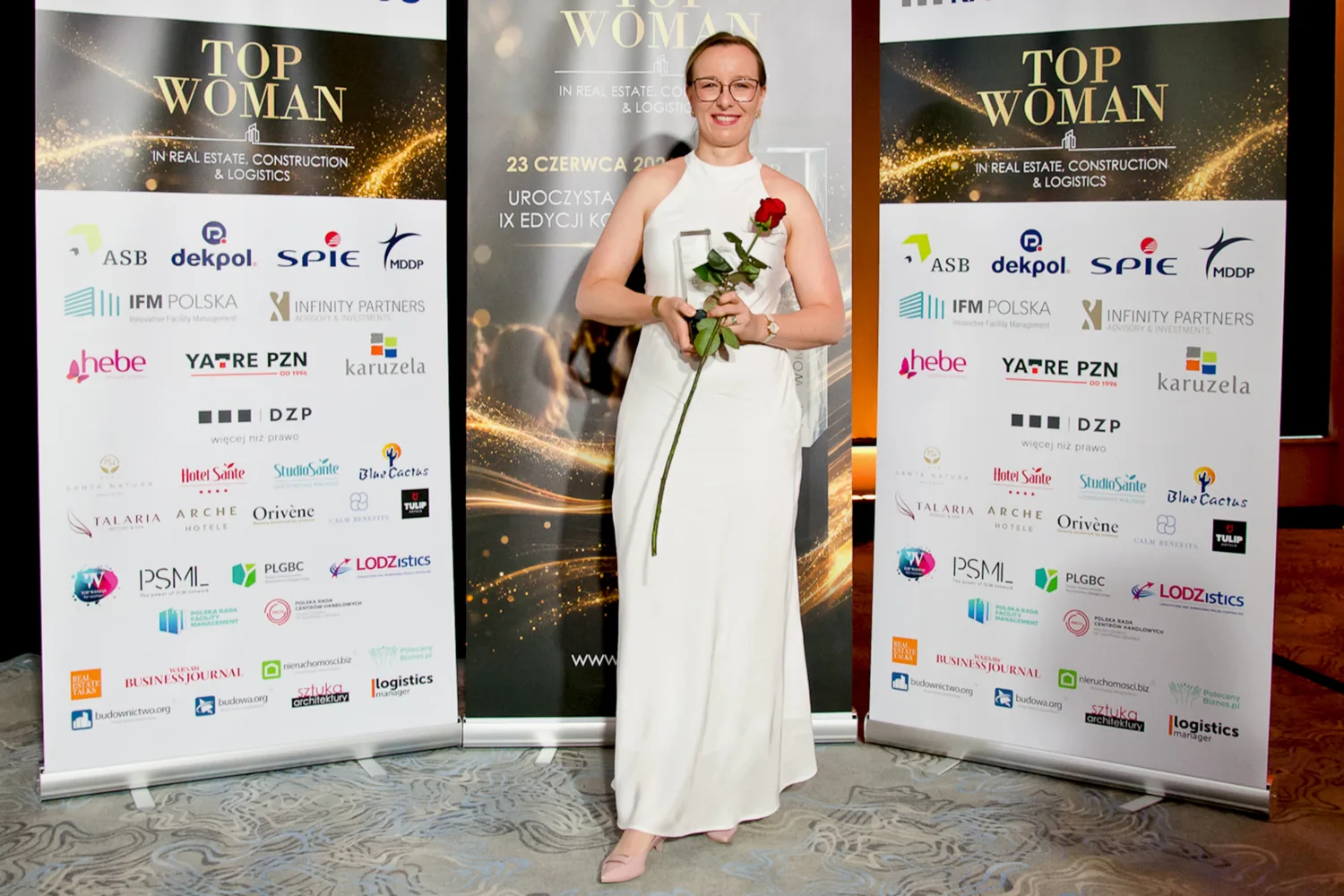 Person holding a glass award and a red rose while standing between event banners displaying sponsor logos and award branding.
