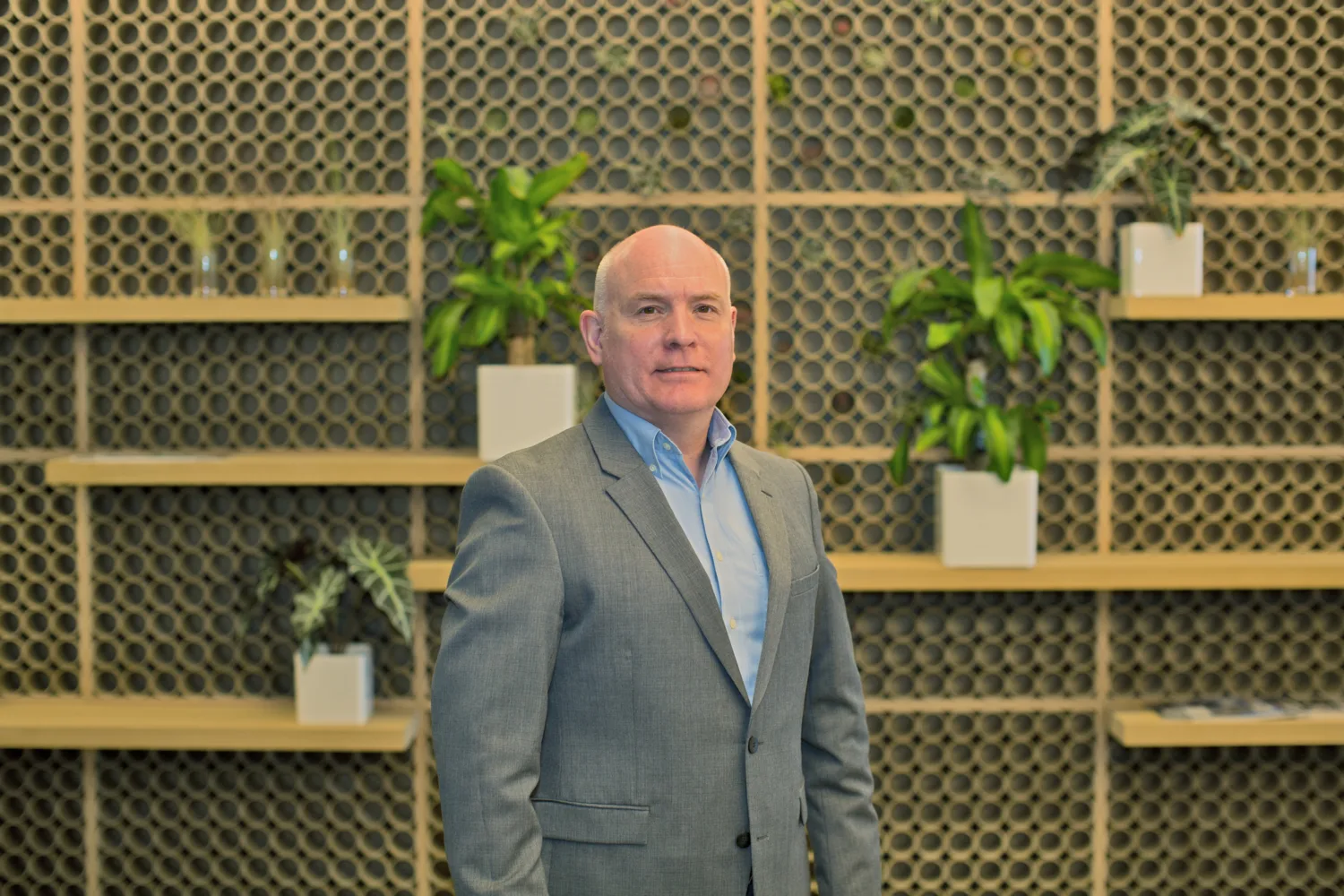Richard Stratton in a pale blue shirt and grey suit jacket against a wooden wall with shelves and plants in white pots