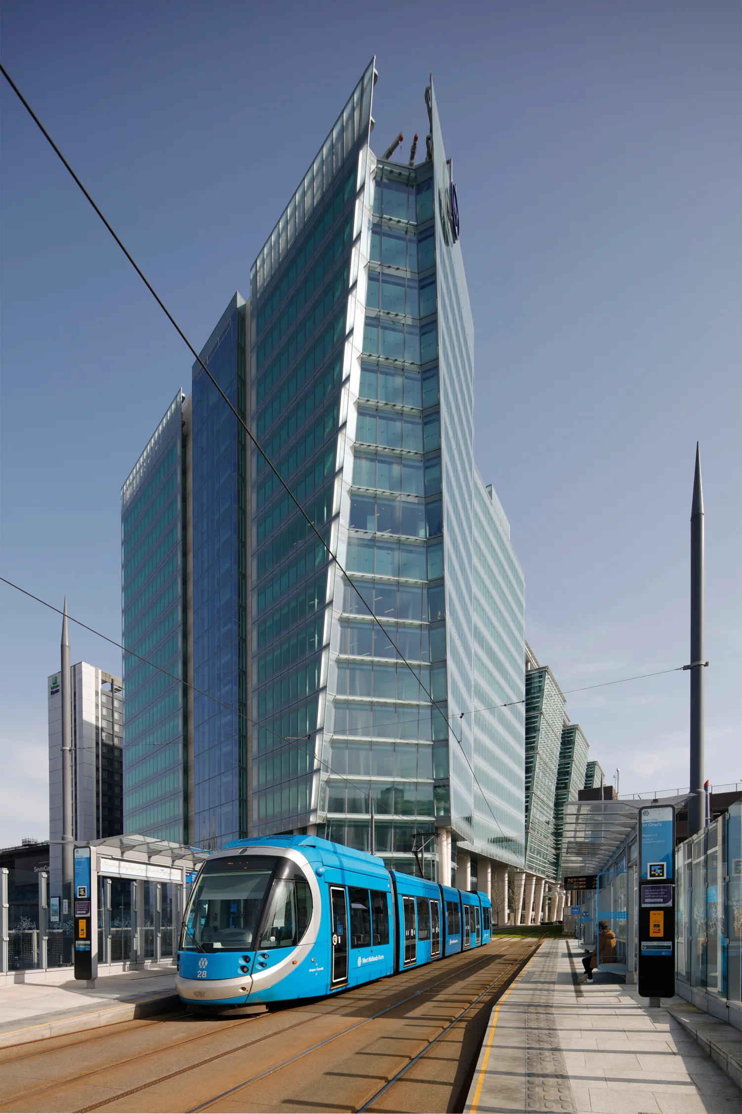 Exterior view of Three Snowhill, Birmingham, a tall windowed building against a blue sky, in the foreground is a blue tram at a tram stop