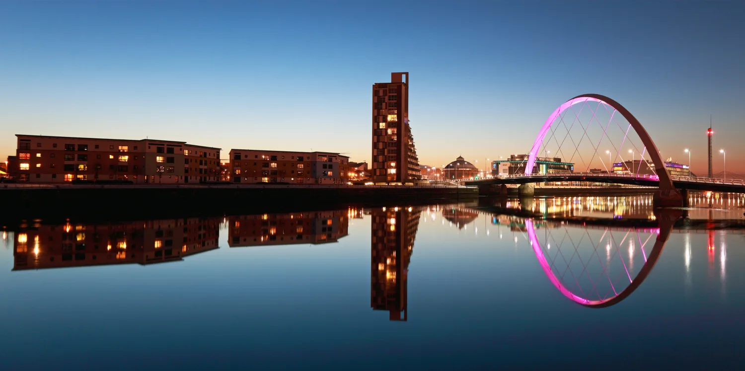Buildings and a pink-lit bridge in Glasgow reflected in the still river