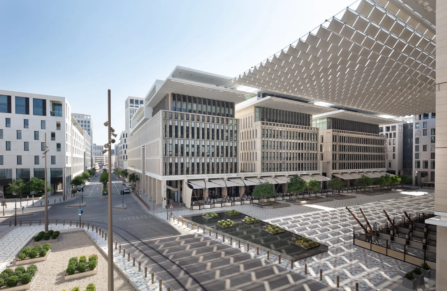 Modern urban plaza with mid-rise buildings featuring glass windows and white facades, patterned pavement, landscaped plant areas, and a large overhead shade structure.