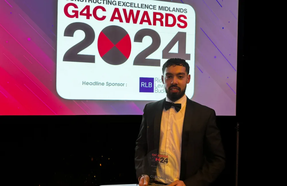 Bilal Arshad wearing a suit holding his award in front of a screen with the G4C Midlands Awards 2024 logo on it