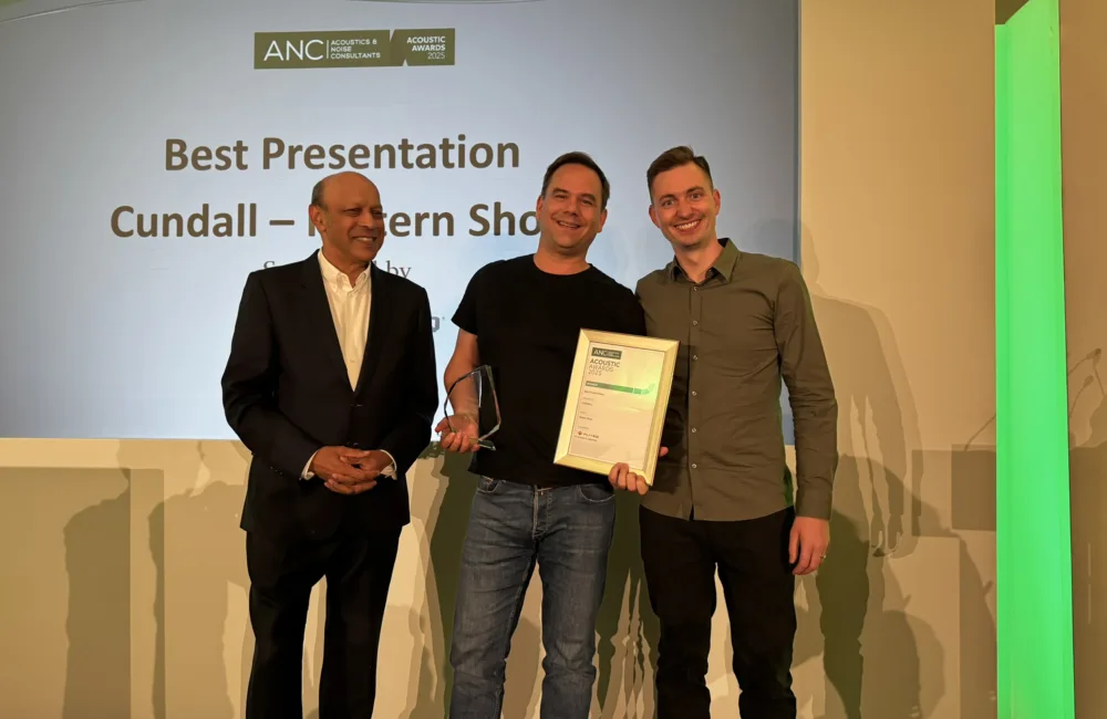 Three people stand on a stage. The person on the left wears a suit; Victor Linstrom holds a glass award and a framed certificate. A screen behind them reads: 'ANC Acoustics & Noise Consultants Association of Noise Consultants Best Presentation Cundall - Pattern Shop.