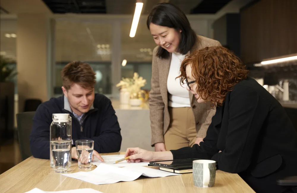 Three people gathered around a wooden table in an office setting, reviewing documents. The table holds papers, a notebook, a ceramic mug, a glass water bottle, and two drinking glasses. Background includes modern cabinetry and decorative plants.