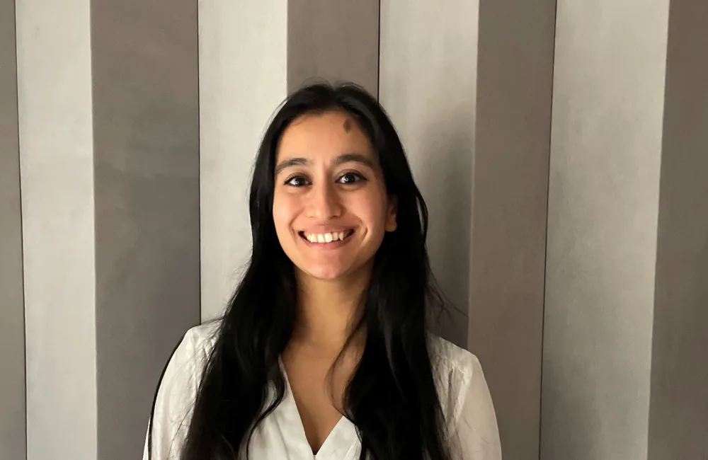 Head-and-shoulders portrait of a person with long dark hair wearing a white top, facing the camera with teeth visible. Background shows vertically striped wall panels in neutral tones; no visible text or additional objects.