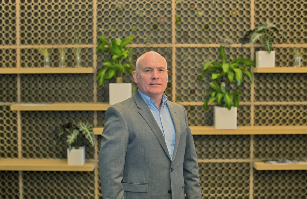 Richard Stratton in a pale blue shirt and grey suit jacket against a wooden wall with shelves and plants in white pots