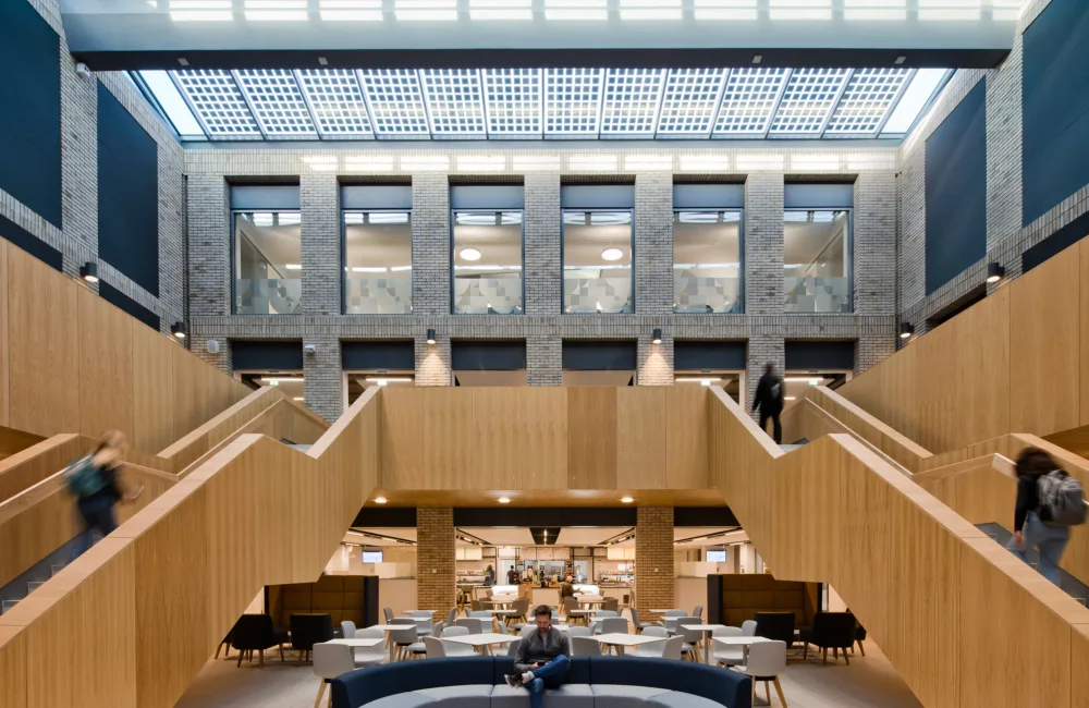 Interior of a university building, large light brown wooden staircase with two sets of stairs at either side of the frame, seating area below the stairs and a grey brick and window floor visible above the stairs
