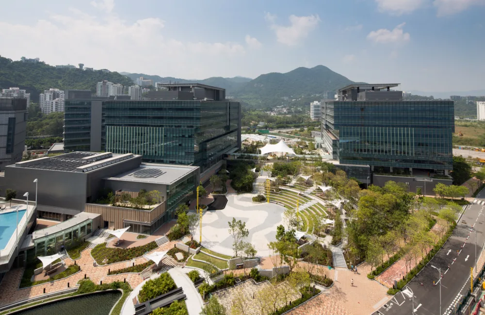 A collection of large modern buildings in a park of cream pathways and vegetation, in the background are green hills and mountains