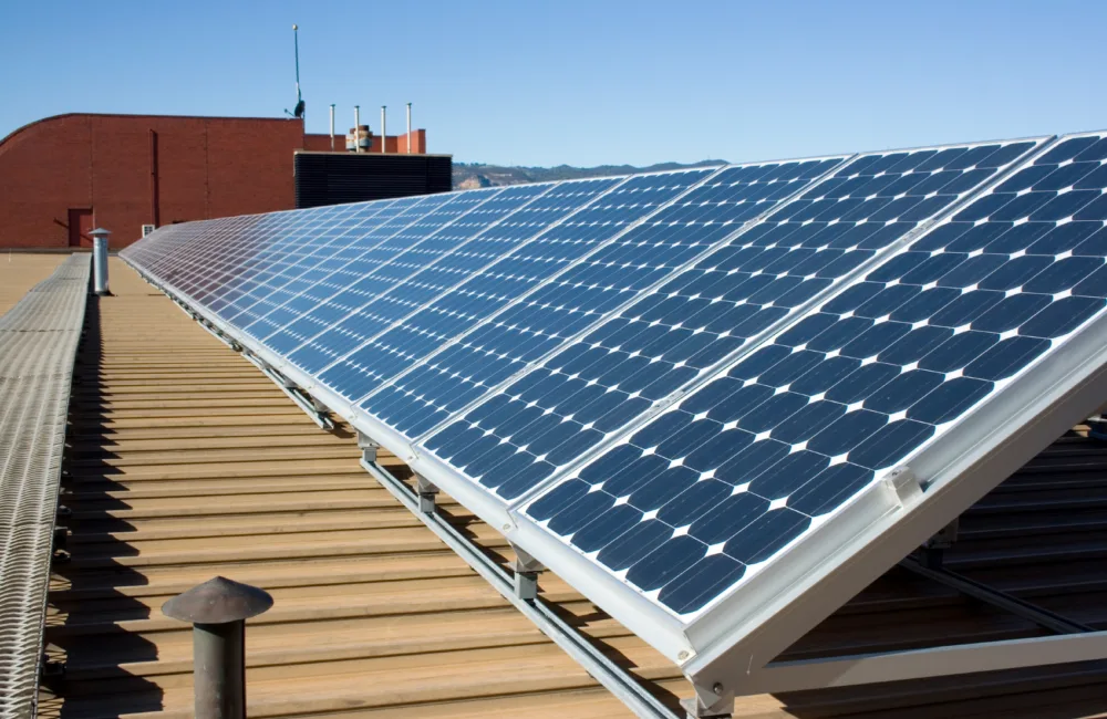Row of angled solar panels on a wood-panelled roof, in the background a red bricked structure is visible