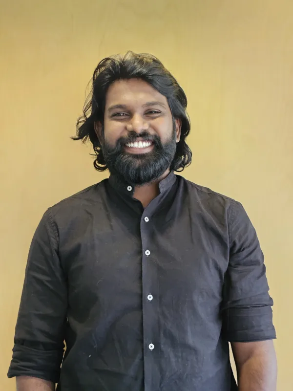 Jotham stands against a plain light‑wood background wearing a dark long‑sleeved shirt with the sleeves rolled up.