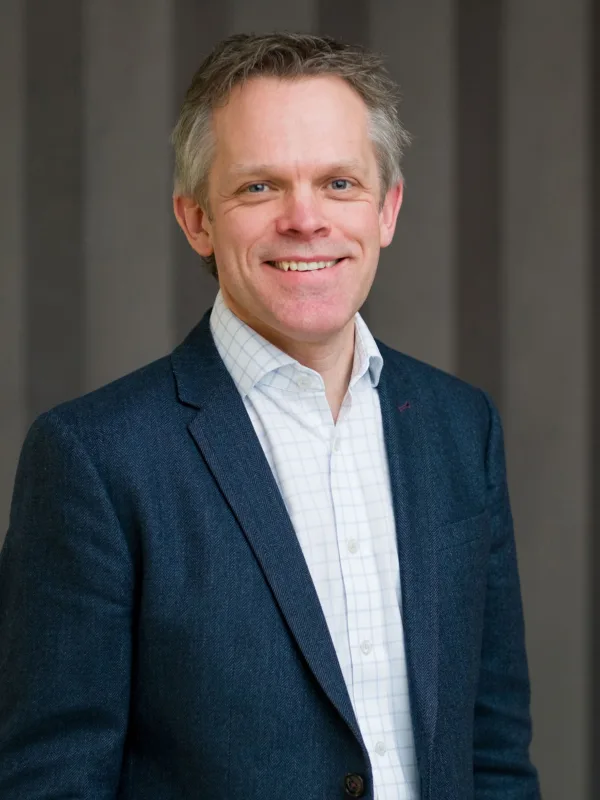 Alex in a dark suit jacket and light open collar shirt, smiling to camera in front of a panelled wall.