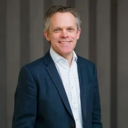 Alex in a dark suit jacket and light open collar shirt, smiling to camera in front of a panelled wall.