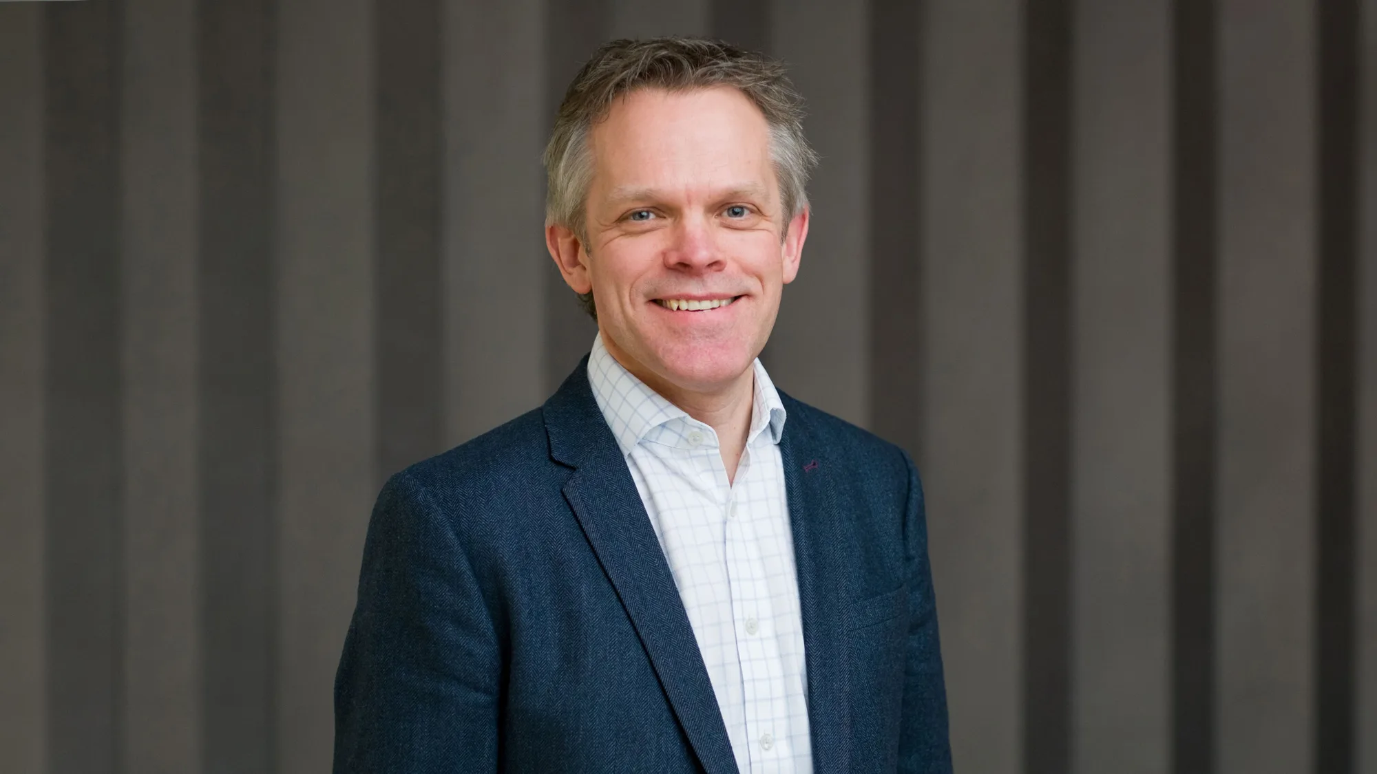 Alex in a dark suit jacket and light open collar shirt, smiling to camera in front of a panelled wall.