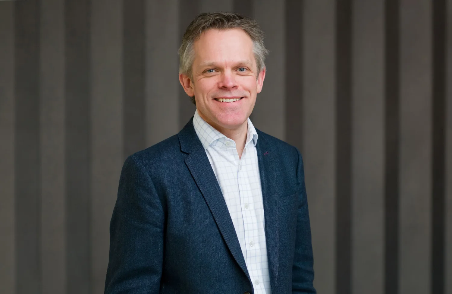 Alex in a dark suit jacket and light open collar shirt, smiling to camera in front of a panelled wall.