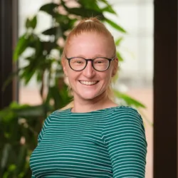 Alison smiling to camera in a green striped long sleeve top in front of a wooden trellis and plant