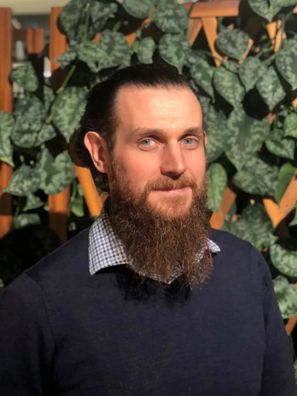 Indoor portrait of a person with long hair tied back and a full beard, wearing a dark sweater over a checkered collar. The background features a wooden lattice partially covered with green leafy plants.