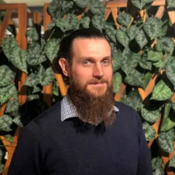 Indoor portrait of a person with long hair tied back and a full beard, wearing a dark sweater over a checkered collar. The background features a wooden lattice partially covered with green leafy plants.