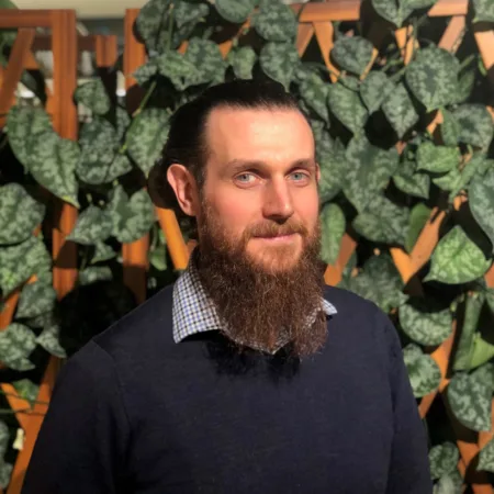 Indoor portrait of a person with long hair tied back and a full beard, wearing a dark sweater over a checkered collar. The background features a wooden lattice partially covered with green leafy plants.