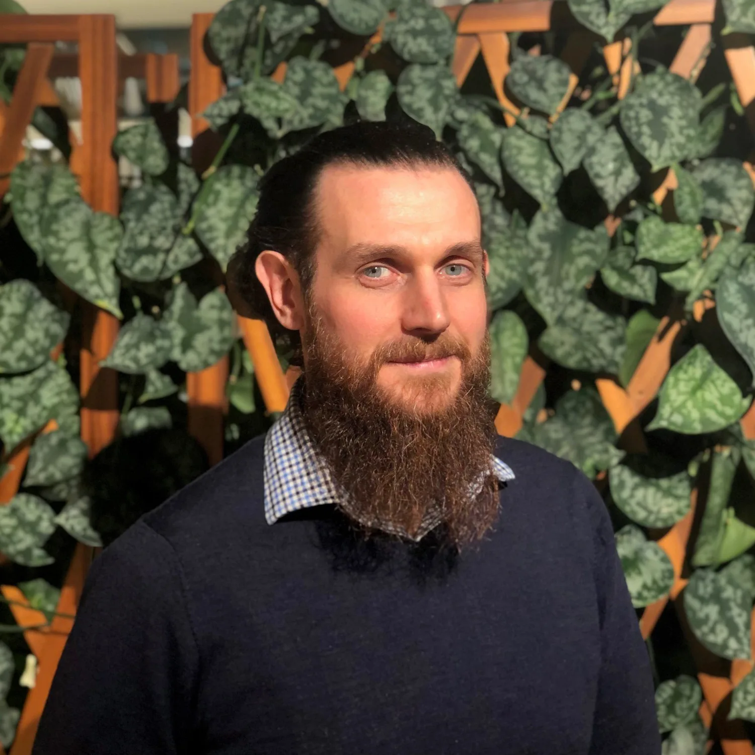 Indoor portrait of a person with long hair tied back and a full beard, wearing a dark sweater over a checkered collar. The background features a wooden lattice partially covered with green leafy plants.