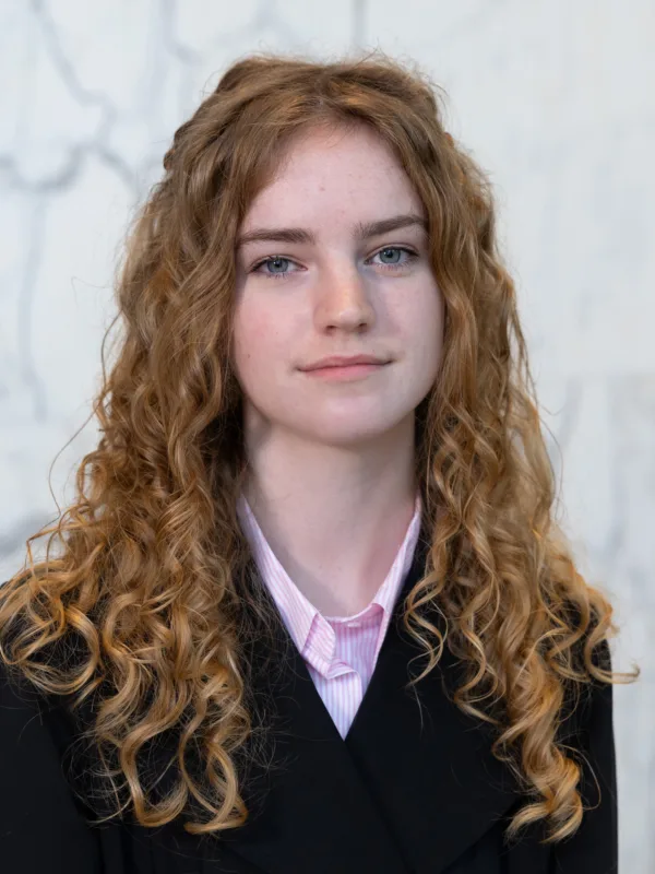 Portrait photograph of a person with long, curly ginger hair wearing a black jacket over a light pink collared shirt, standing against a pale grey and white marble wall background.