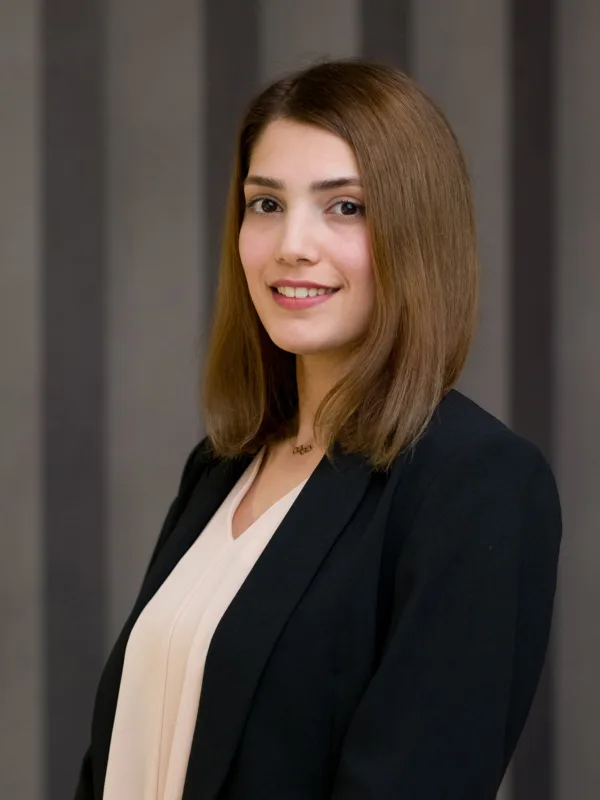 Studio-style portrait of a person standing three-quarters to the camera, wearing a dark blazer over a light blouse, with shoulder-length straight hair, set against a neutral grey curtain backdrop.
