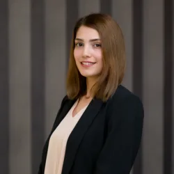 Studio-style portrait of a person standing three-quarters to the camera, wearing a dark blazer over a light blouse, with shoulder-length straight hair, set against a neutral grey curtain backdrop.