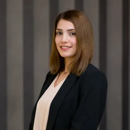 Studio-style portrait of a person standing three-quarters to the camera, wearing a dark blazer over a light blouse, with shoulder-length straight hair, set against a neutral grey curtain backdrop.