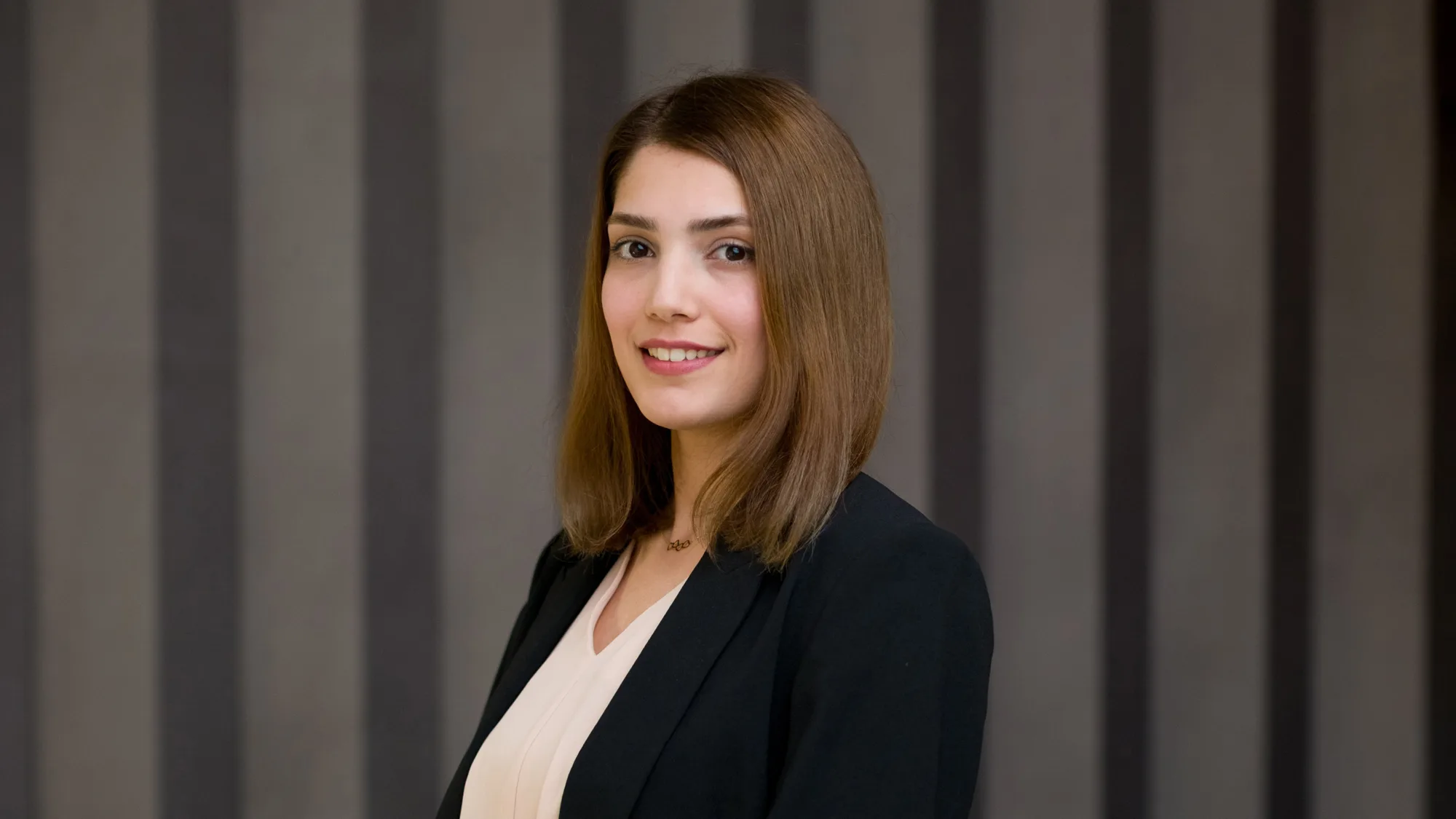 Studio-style portrait of a person standing three-quarters to the camera, wearing a dark blazer over a light blouse, with shoulder-length straight hair, set against a neutral grey curtain backdrop.