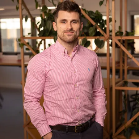 Ian stood with hands in pockets in a pink long sleeve shirt in front of a plant trellis in an office