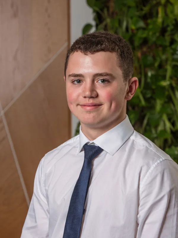 Person wearing a white dress shirt and dark patterned tie is seated indoors. Background features a wooden panel with diagonal lines on the left and a green vertical plant wall on the right.
