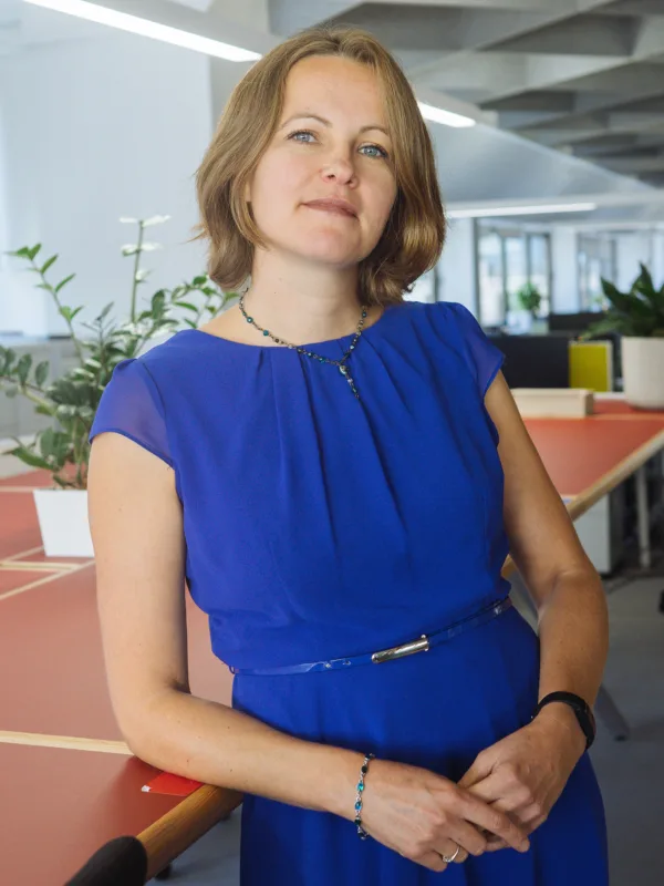 A person in a blue dress stands beside a large shared workspace table in a modern, open‑plan office with plants, shelving, and desks visible in the background.