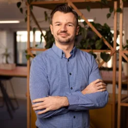 Maciej stood in a blue shirt with arms folded in front of a wooden shelving structure