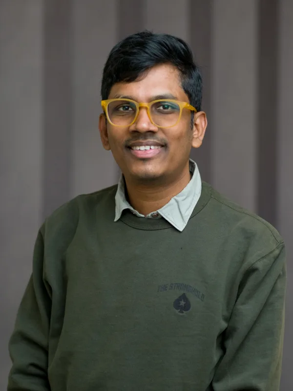 Person wearing a dark green crew-neck sweatshirt with a small emblem on the left chest and a light grey collared shirt underneath, seated against a background of vertical dark grey panels.