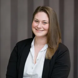 A person with shoulder-length brown hair, wearing a white button-up shirt and black blazer, seated in front of a background with vertical grey stripes.