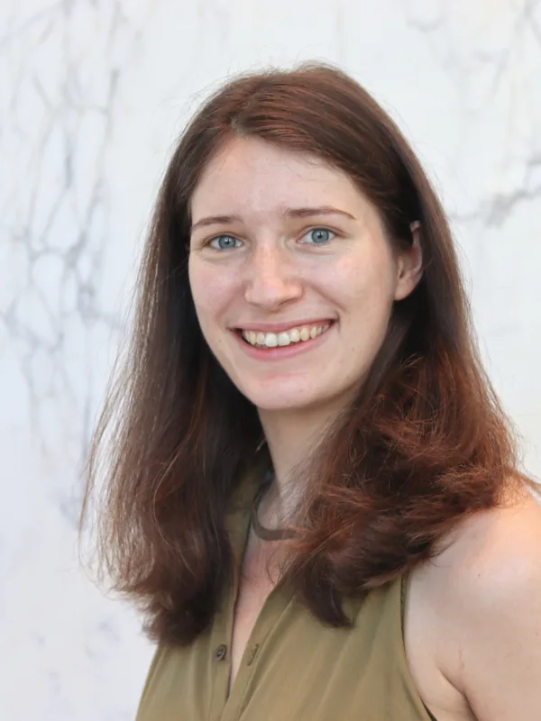Head-and-shoulders portrait of a person with shoulder-length reddish-brown hair, facing the camera with teeth visible, wearing a sleeveless olive-green top, standing against a light marble-patterned wall.