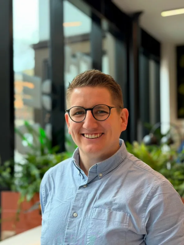 A person wearing round glasses and a light blue button-up shirt is shown from the chest up indoors, standing near large windows with black frames, with potted plants and a blurred cityscape visible in the background.