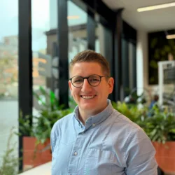 A person wearing round glasses and a light blue button-up shirt is shown from the chest up indoors, standing near large windows with black frames, with potted plants and a blurred cityscape visible in the background.