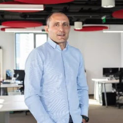 Horia stood wearing a blue open collared shirt stood in an open plan office