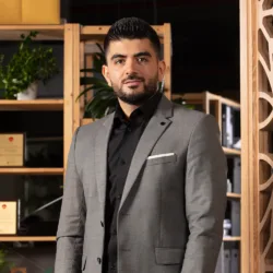 Mahmoud in a grey suit jacket and black shirt looking to camera in front of an in interior wooden trellis and shelving unit