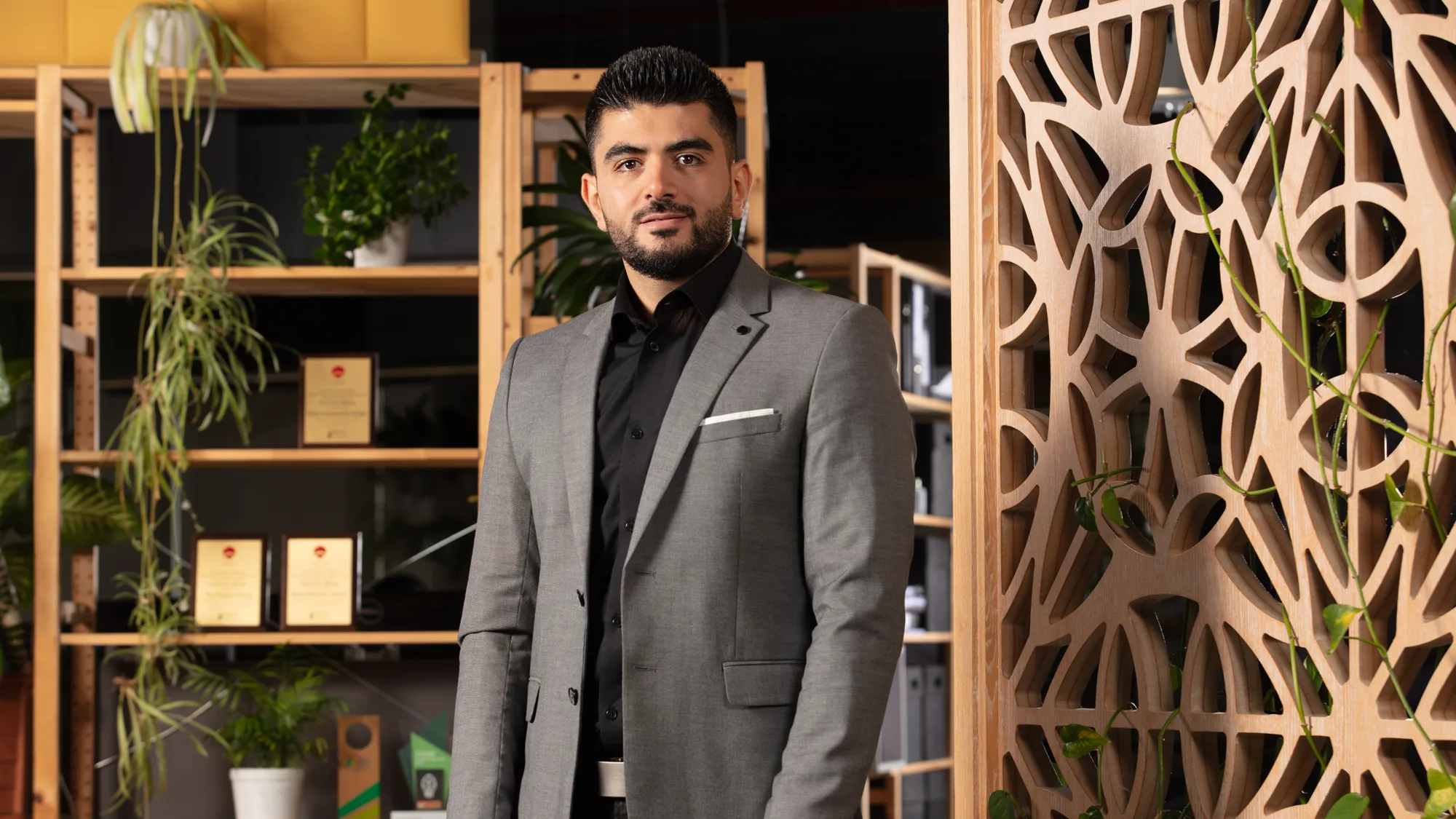 Mahmoud in a grey suit jacket and black shirt looking to camera in front of an in interior wooden trellis and shelving unit