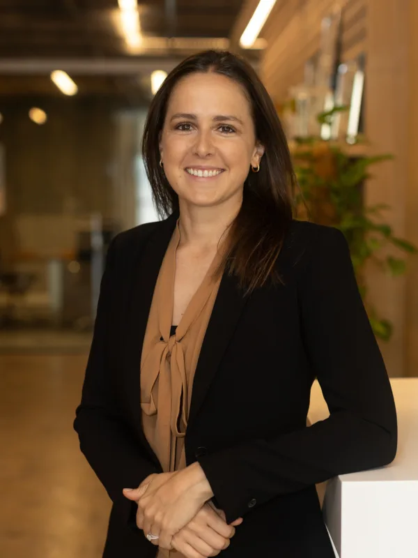 Annabelle standing indoors wearing a black blazer over a brown blouse. The background shows an office with glass walls, ceiling lights, and a plant on the right side.